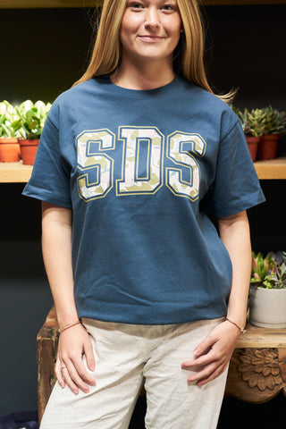 Indoors, someone wears the SDS Prep Fruit Fields Short Sleeve Tee featuring bold “SDS” letters, paired with light pants. Shelves of potted plants in the background add a cozy touch to this exclusive Bravocon setting.