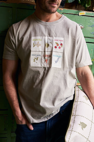 A man wearing the Heirloom Seeds Short Sleeve Tee, featuring seed packet illustrations—Green Pea, Cherry Tomato, Sweet Basil, Carrot, and Rosemary—stands by a rustic green cabinet with his hand in his pocket.