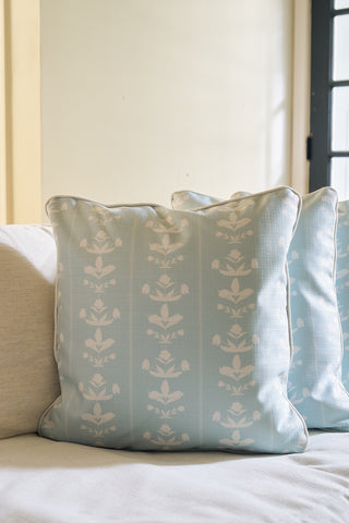 Two Garden Party Pillows in light blue with white floral patterns from The Core Collection rest on a light-colored sofa near a window, adding a cozy and inviting touch to the space.