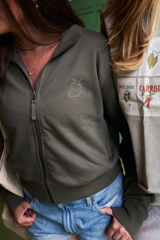 Two women stand close together. One wears the Very Berry Embroidered Full-Zip Jacket with a small strawberry outline and blue jeans; the other sports a Storyteller Embroidered Crewneck Sweatshirt with sweet basil and carrot seed packets. Their faces are hidden.