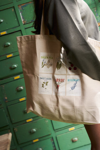 A person with an Heirloom Seeds Tote Bag featuring botanical prints stands before green lockers, sporting a Bravocon exclusive Craig sweatshirt that matches their effortlessly cool style.