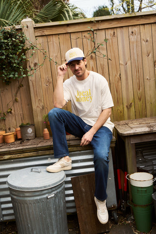 A man wearing a Local Honey Embroidered Hat sits on a wooden bench in a backyard garden, surrounded by potted plants, gardening tools, and a trash can against a wooden fence backdrop.