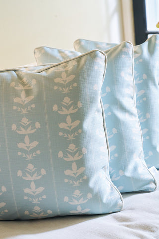 Three light blue Garden Party Pillows with white floral covers are arranged in a row on a light surface, sunlight streaming in from a nearby window.
