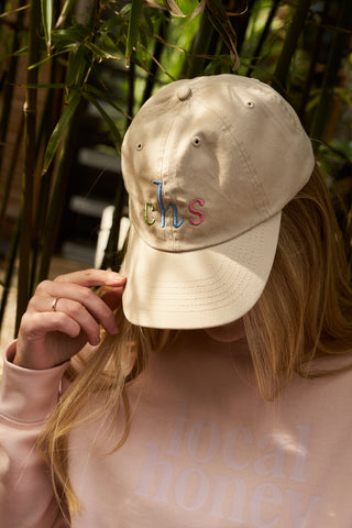 A person with long blonde hair sits outdoors near bamboo, their face partially hidden by a Rainbow Row Dad Cap embroidered with colorful "chs" letters, and they wear a light pink "local honey" sweatshirt.