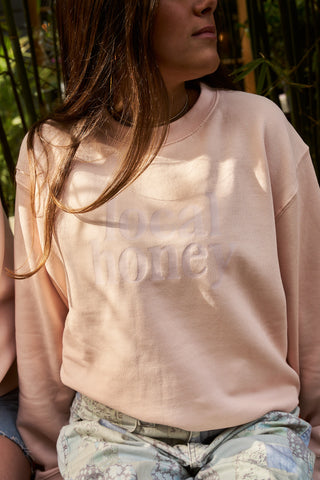 A person with long brown hair wears a light pink cotton blend sweatshirt, like the Local Honey Crewneck Sweatshirt, paired with light patterned pants while seated outdoors.