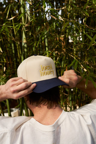 A person adjusts the brim of their Local Honey Embroidered Hat while facing away from the camera, surrounded by tall green bamboo.