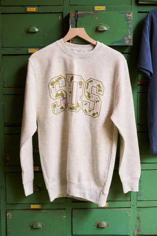 A light beige SDS Prep Fruit Fields Crewneck Sweatshirt with large camouflage SOS letters on the front hangs on a wooden hanger in front of vintage green lockers.