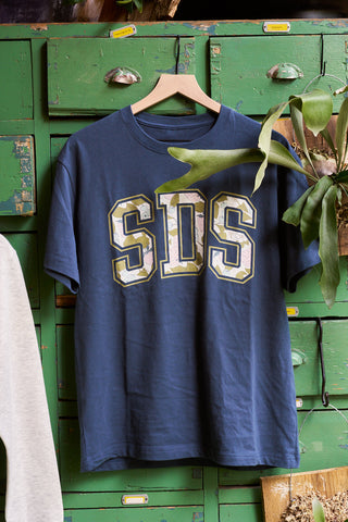 The SDS Prep Fruit Fields Short Sleeve Tee, a blue cotton blend with bold "SDS" block letters, hangs on a wooden hanger in front of green vintage lockers while a leafy plant drapes over part of the shirt.