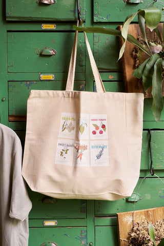 The Heirloom Seeds Tote Bag, featuring illustrations and names of green pea, cherry tomato, sweet basil, carrot, and rosemary, hangs on a vintage green cabinet—ideal for carrying your favorite cotton blend sweatshirt.