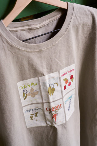 The exclusive Heirloom Seeds Short Sleeve Tee in beige cotton blend hangs on a wooden hanger, displaying six illustrated seed packets—Green Pea, Cherry Tomato, Sweet Basil, Carrot, and Rosemary—each with charming plant drawings.
