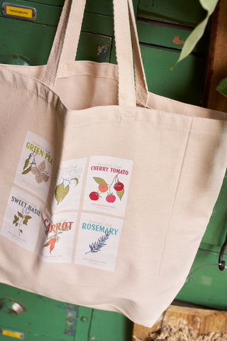 The Heirloom Seeds Tote Bag, in beige canvas, hangs on a door and features illustrated seed packets for green pea, jalapeño, cherry tomato, sweet basil, carrot, and rosemary—a charming Bravocon exclusive.
