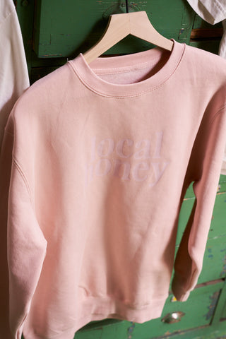 The Local Honey Crewneck Sweatshirt in light pink, featuring "local honey" subtly printed in white, is displayed on a wooden hanger against a green background.