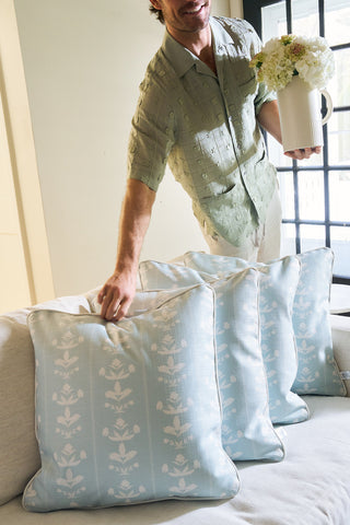A person in a light green shirt touches the Garden Party Pillow, a blue and white floral cover, while holding a white pitcher of white flowers. Sunlight streams through the window behind them.