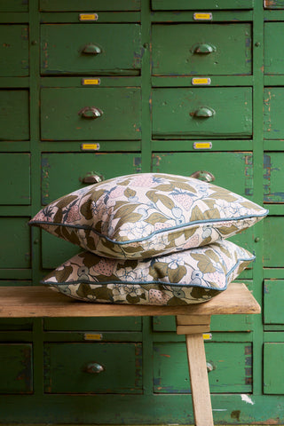 Two Fruit Fields Pillows with pink and green fruit and floral covers are stacked on a wooden bench, bringing fresh garden charm in front of a vintage green cabinet with yellow label holders.