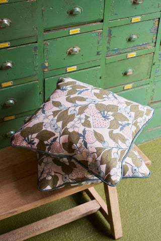 Two Fruit Fields Pillows with pink, green, and blue floral patterns are stacked on a wooden bench. Behind them, a vintage green cabinet with small drawers and yellow labels completes the charming garden decor scene.