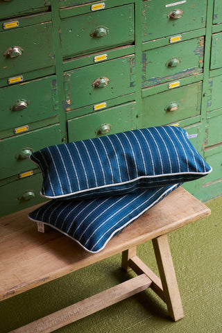Two Courtyard Stripes Extended Lumbar Pillows are stacked on a wooden bench in front of green vintage filing cabinets with metal handles, set atop a green carpeted floor.