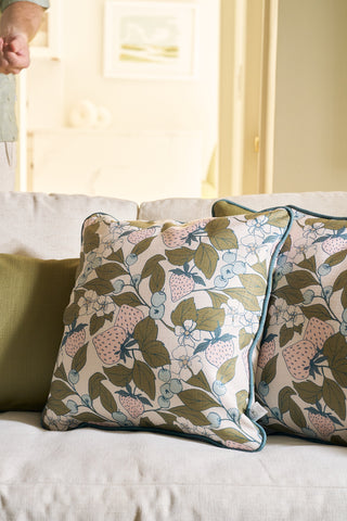 Two Fruit Fields Pillows with green and pink floral covers sit on a light sofa beside a solid green cushion. A person’s arm is partially visible, art hangs on the wall. Fruit Fields Pillow adds a fresh touch—perfect for garden-inspired decor.