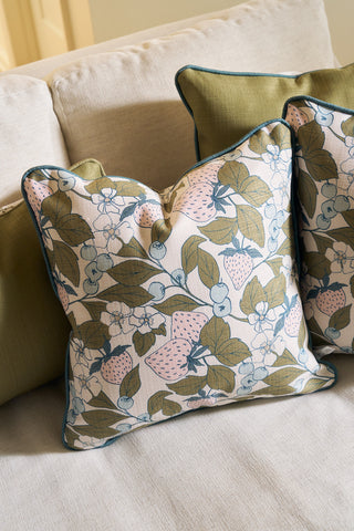Two Fruit Fields Pillows, featuring a botanical strawberry and leaf print, rest on a beige sofa with a green pillow in the background. These pillow covers have piped edges and offer a soft, inviting look.