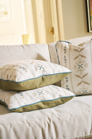 Three decorative pillows, including the Grandmillenial Garden Pillow with green edging, rest on a light-colored sofa in a cozy, sunlit room. Behind them are a door and framed Core Collection artwork on the wall.