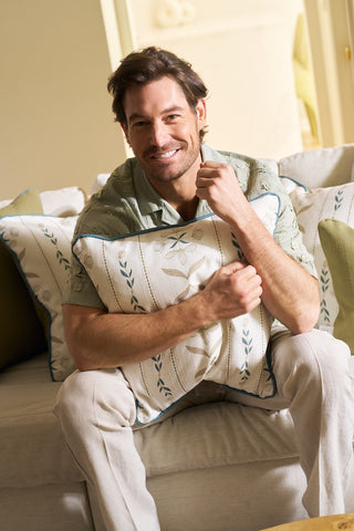 A man with brown hair and a light green shirt smiles on a sofa, hugging the Grandmillenial Garden Pillow. The softly lit living room in neutral tones reflects the Broad Street Blooms style.