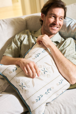 A smiling man in a light green shirt sits on a light-colored couch, holding the Grandmillenial Garden Pillow from the Core Collection, which features a soft blue and beige leaf pattern.