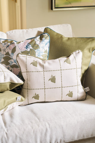 A beige sofa features several decorative pillows, including a Broad Street Blooms floral cover, the Strawberry Stitch Grid Lumbar Pillow with a green grid and strawberry pattern, and a solid green Core Collection pillow in the background.