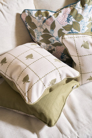 Three decorative pillows from the Core Collection on a cream couch: a Broad Street Blooms pillow cover with green and pink fruit, a Strawberry Stitch Grid Lumbar Pillow in green plaid with strawberries, and one solid olive green pillow.