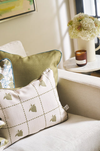 A beige sofa from the Core Collection with decorative pillows, including a Strawberry Stitch Grid Lumbar Pillow, sits beside a small round table holding a vase of white flowers and a lit brown candle.