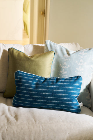 Three decorative pillows from the Core Collection rest on a light beige couch: a green square pillow, a light blue floral pillow cover, and the Courtyard Stripes Extended Lumbar Pillow. A softly lit room appears in the background.