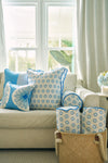 A light beige sofa with blue and white patterned pillows, including a Calico Mini Floral Oversized Pillow with bright blue piping, sits by large windows with sheer curtains. A woven basket holds matching bolsters; a vase of blue flowers is on a side table.