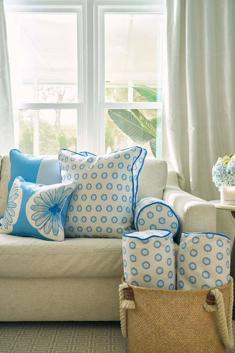 A light beige sofa with blue and white patterned pillows, including a Calico Mini Floral Oversized Pillow with bright blue piping, sits by large windows with sheer curtains. A woven basket holds matching bolsters; a vase of blue flowers is on a side table.