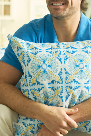 A man in a blue shirt hugs the Euro Blue Tiles Pillow, featuring a blue, yellow, and white floral design. Only the lower half of his smiling face is visible as the pillow adds charm to the scene.