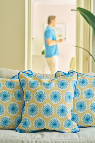 The Yellow Calico Scallops Pillow, with blue floral patterns and piping, creates a sophisticated accent on a beige sofa. In the blurred background, a person in a blue shirt stands holding a cup.