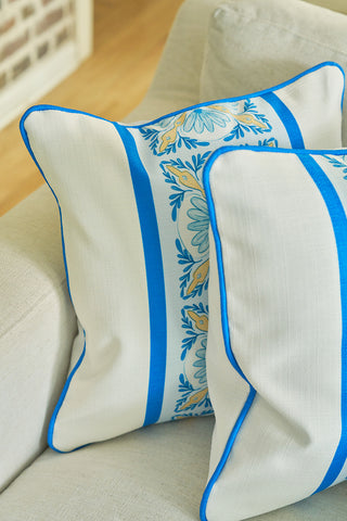 Two cream cushions rest on a light sofa; one is the Line of Euro Blue Tiles Pillow, featuring a central blue and yellow floral pattern with bold blue stripes, bringing subtle European elegance and a coastal touch.