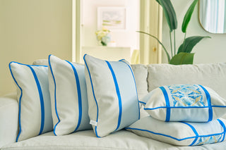 Three decorative blue-striped pillows and a Line of Euro Blue Tiles Pillow with floral embroidery are arranged on a light sofa, adding European elegance. A leafy plant and framed art accent the bright, softly lit background.