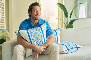 A man in a blue shirt sits on a light sofa, smiling and holding a Line of Euro Blue Tiles Pillow that adds European elegance. Another matching pillow is beside him amid plants, flowers, and natural light for a coastal vibe.