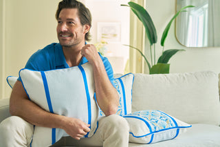 A man in a blue shirt smiles while sitting on a light-colored couch, hugging a Line of Euro Blue Tiles Pillow. More pillows and a large plant in the background add European elegance to this cozy, bright living room.