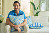 A man with light brown hair and a blue shirt sits on a beige couch, smiling and hugging a blue and white floral pillow. Beside him is the Euro Blue Tiles with Spring Blue Stripe Pillow, adding European elegance to the space.