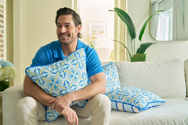 A man with brown hair in a blue shirt and beige pants sits on a light sofa, hugging the Euro Blue Tiles with Spring Blue Stripe Pillow. Coordinated pillows and green plants lend European elegance to the bright living room.
