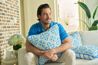 A man with brown hair and a beard, in a blue shirt and light pants, sits on a white sofa hugging the Spring Blue Stripe Pillow from the Euro Blue Tiles collection. Patterned pillows, flowers, and a brick wall evoke European elegance in the background.