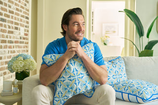 A man with brown hair and a beard smiles on a white couch, holding a Spring Blue Stripe Pillow. The room features Euro Blue Tiles, plants, flowers, soft blue accents, and subtle European elegance.