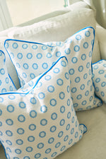 Three white throw pillows with blue sunburst designs and bright blue piping rest on a light sofa. One is the Calico Mini Floral Oversized Pillow, featuring a subtle mini floral pattern. Sunlight streams through the nearby window, brightening the scene.