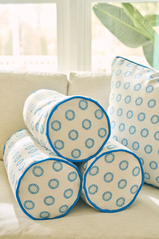 Three Calico Mini Floral Bolster Pillows featuring vibrant blue flowers are stacked on a light-colored couch, with a matching square pillow in the background near a leafy plant as natural light fills the room.