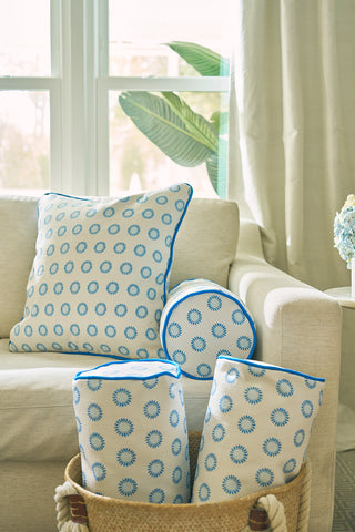 A beige sofa with blue and white patterned pillows and the Calico Mini Floral Bolster Pillow sits by a bright window. Matching pillows fill a basket on the floor, while light curtains, a green plant, and vibrant blue flowers add to the cozy look.