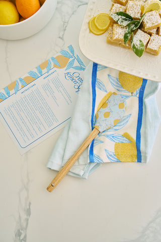 On a marble countertop, a recipe card sits beside a bowl of lemons. The heat-resistant Lots of Lemons Spatula, made from BPA-free silicone, rests on a lemon-themed towel. Nearby, lemon bars garnished with lemon slices and mint sit on a plate.