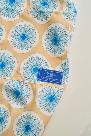 A close-up of fabric displays a pattern of blue flowers on beige circles. A blue label reads Sewing Down South, featuring sewing tools and Est. 2018. The slightly crumpled fabric highlights the charm of an adjustable neck strap Calico Scallops on Yellow Apron against a white background.