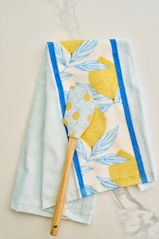 The Lots of Lemons Spatula, made from heat-resistant BPA-free silicone, sits stylishly on a white marble countertop with a plain light blue towel and another adorned with yellow lemons and blue leaves.