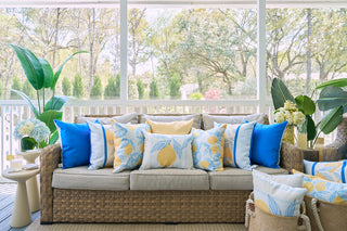 A wicker sofa creates a cozy sun porch nook, accented by the blue, white, and yellow Lots of Lemons Pillow for a vibrant look. Surrounded by lush plants and large windows, it offers views of a garden filled with trees.