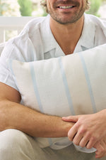 A man in a short-sleeved white shirt smiles while holding the Lemons in a Line Lumbar Pillow, featuring subtle blue stripes. The image shows only his upper body and arms.