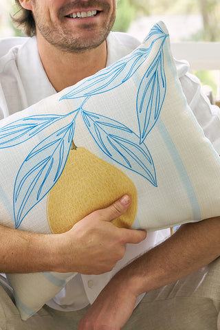 A man smiles while hugging the Lemons in a Line Lumbar Pillow, featuring blue leaf and yellow fruit designs. He wears a white shirt and beige pants, sitting in a bright setting with citrus-inspired decor.